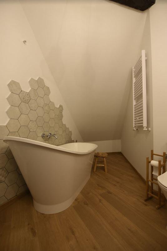 Biała wanna wolnostojąca z baterią ścienną na tle ściany wyłożonej heksagonalnymi płytkami w odcieniach szarości, drewniana podłoga ułożona w jodełkę, drewniany stołek i grzejnik drabinkowy...