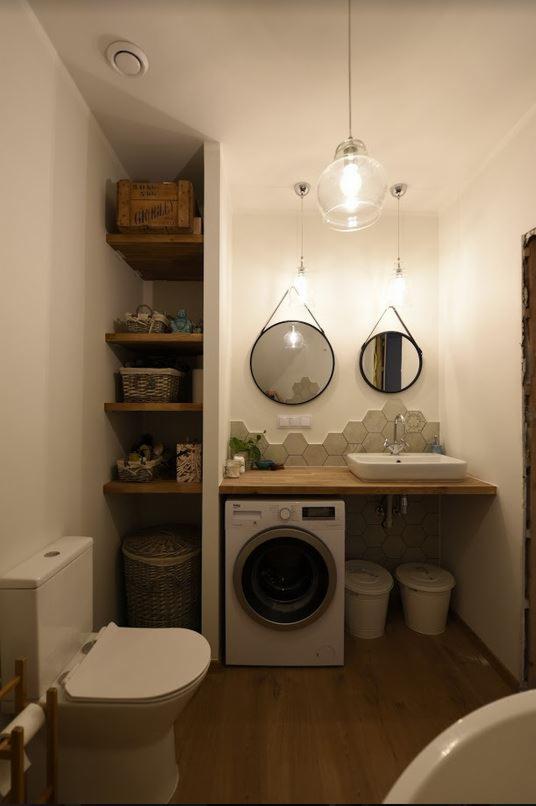 Mała łazienka z pralką pod blatem, heksagonalne płytki, okrągłe lustra na paskach i drewniane półki z koszami