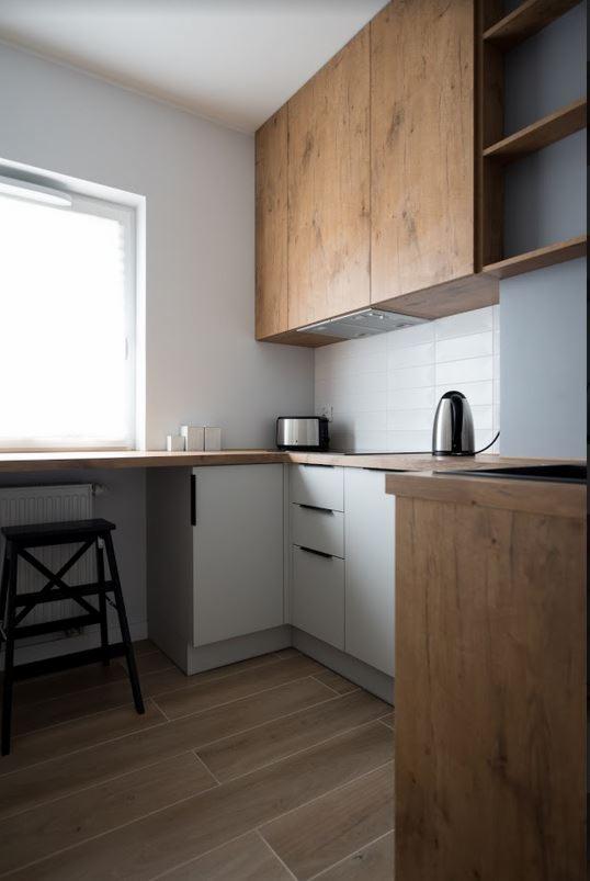 Nowoczesna kuchnia w odcieniach szarości i drewna z drewnianymi szafkami wiszącymi, białymi płytkami na ścianie, srebrnym czajnikiem i tosterem oraz czarnym stołkiem drabinkowym.