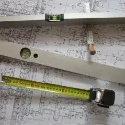 Kątownik, poziomica i zwinięta miara na tle architektonicznego planu budynku.