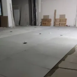 Układanie jasnych płyt podłogowych w dużym, surowym pomieszczeniu z widocznymi otworami na instalacje, paletami w tle i niedokończonymi ścianami.