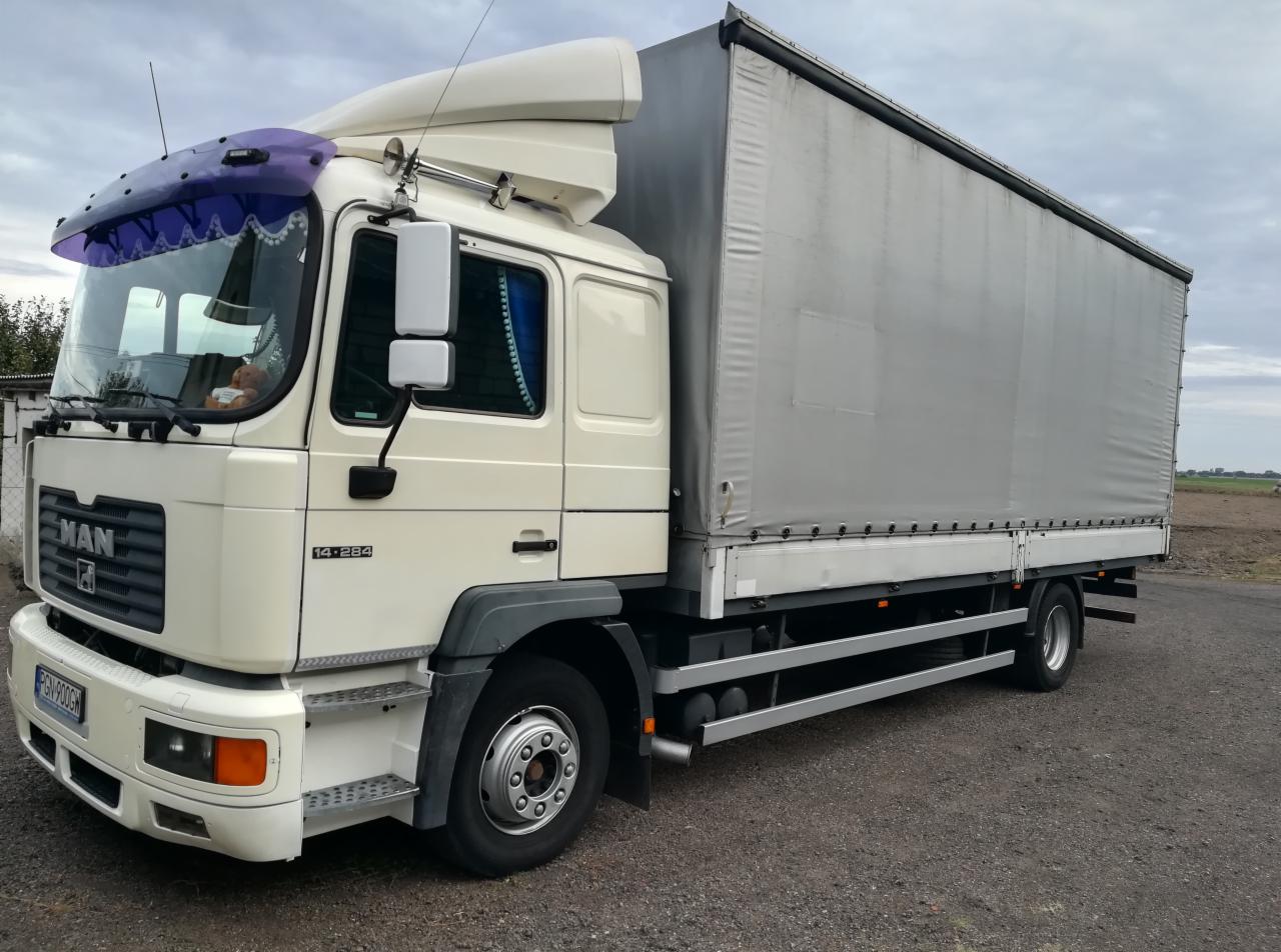 Biała ciężarówka MAN 14.284 z szarą plandeką, zaparkowana na żwirowym podłożu, widoczna z boku pod kątem, z pluszowym misiem za szybą.