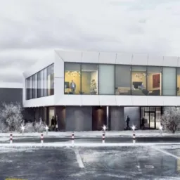 Dwukondygnacyjny budynek biurowy z dużymi oknami, widoczne biura wewnątrz, zimowa sceneria, zaparkowane samochody i maszty z flagami firmy.