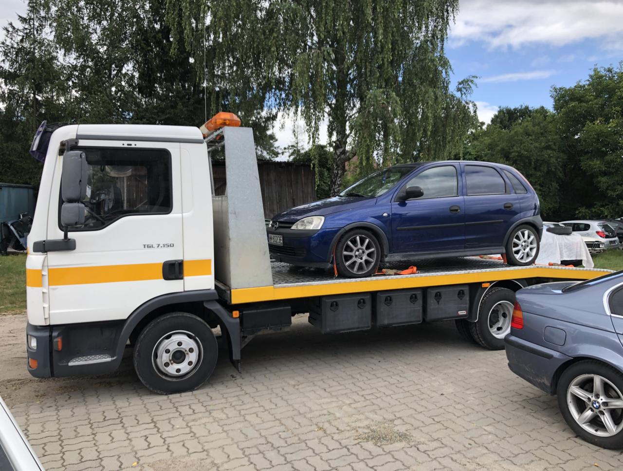 Biała ciężarówka transportowa MAN TGL 7.150 z załadowanym na żółtej platformie niebieskim samochodem osobowym Opel Corsa na brukowanym placu, z widocznymi drzewami w tle.