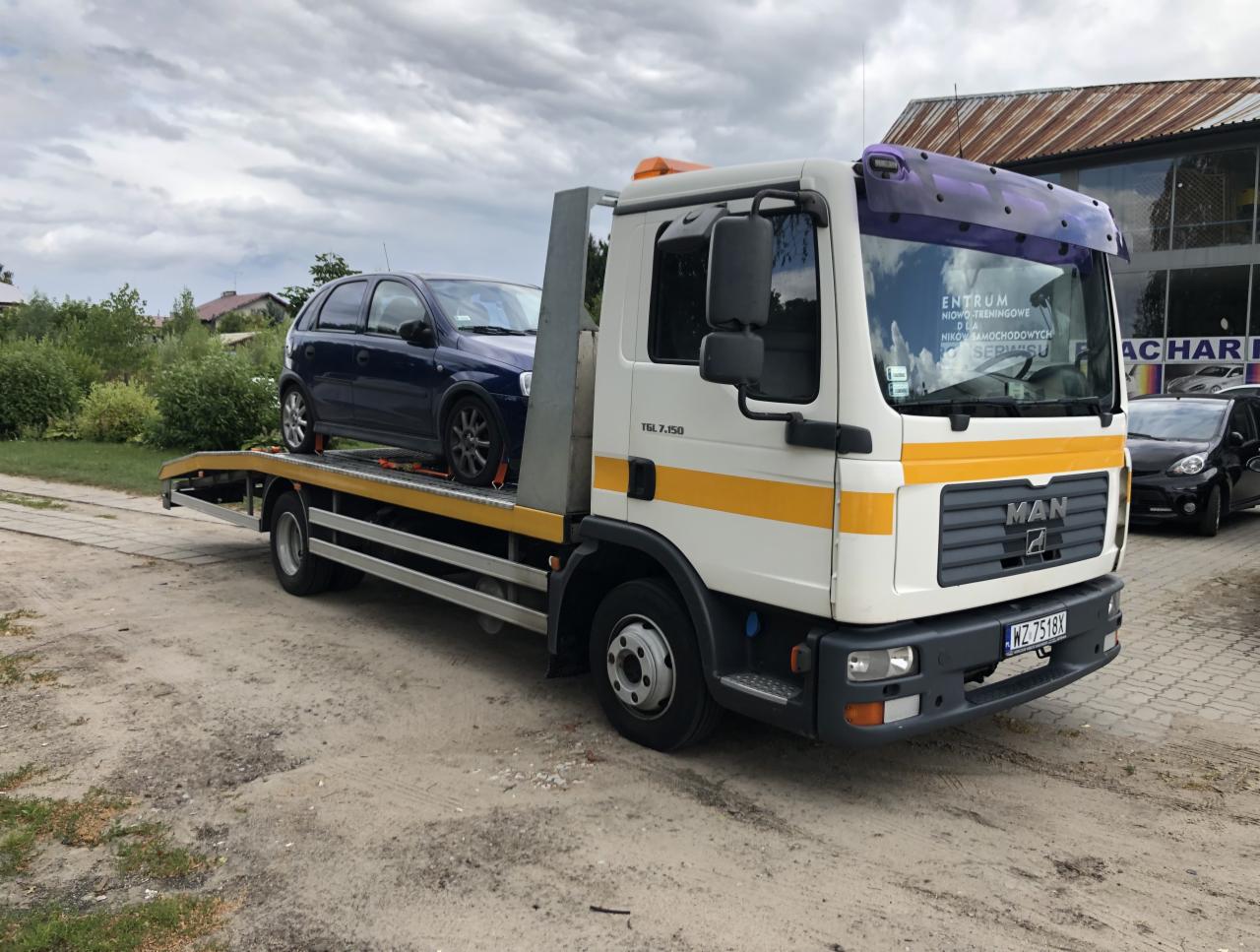 Biała laweta MAN TGL 7.150 z żółtymi pasami transportuje niebieski samochód osobowy Opel Corsa. W tle budynek serwisu samochodowego i drzewa.