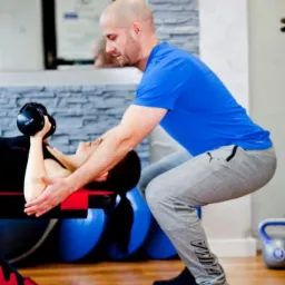 Mężczyzna w niebieskiej koszulce asekuruje kobietę ćwiczącą z hantlami na ławce w siłowni, w tle lustro i sprzęt do ćwiczeń.