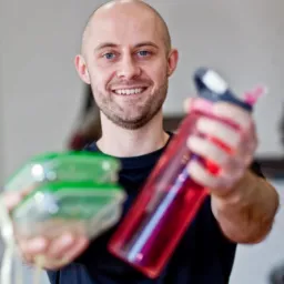 Uśmiechnięty mężczyzna z krótkimi włosami prezentuje pojemnik z jedzeniem i czerwoną butelkę na wodę, w tle fragment drążka do ćwiczeń.