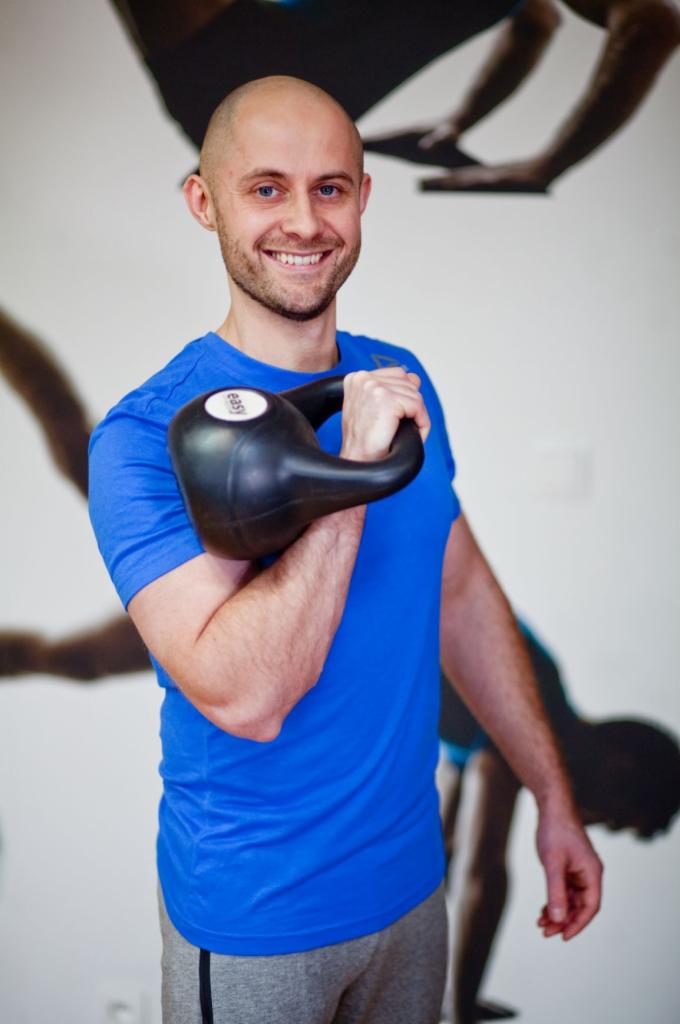 Uśmiechnięty mężczyzna w niebieskiej koszulce trzyma kettlebell na ramieniu, w tle grafika przedstawiająca sylwetki ćwiczących osób.