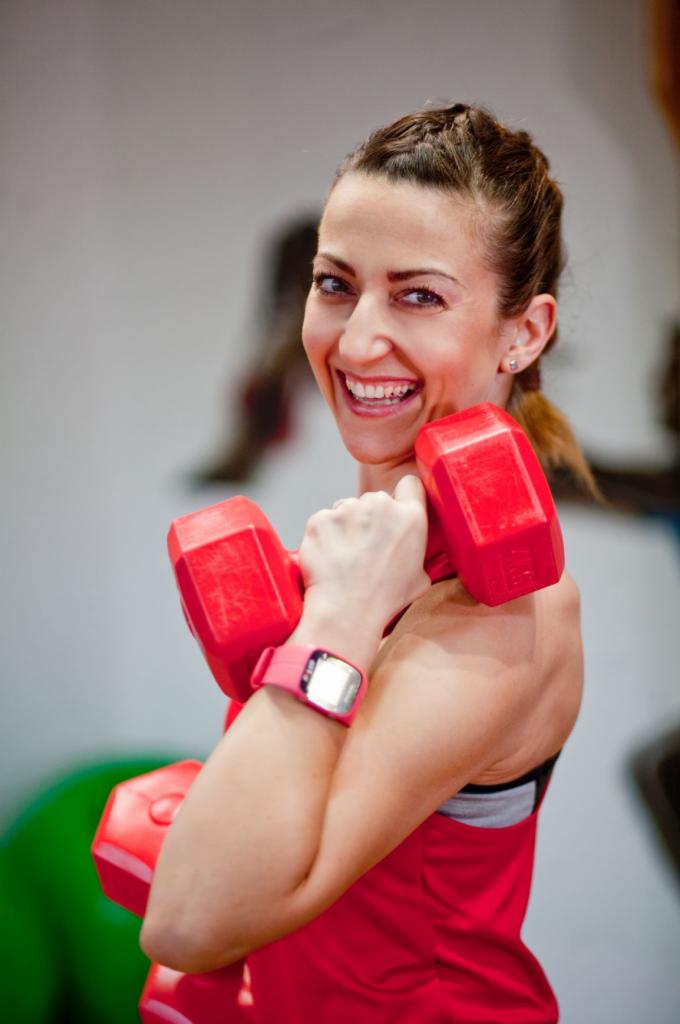Uśmiechnięta kobieta w czerwonej koszulce trzyma czerwone hantle, na nadgarstku różowy smartwatch, w tle sprzęt do ćwiczeń.