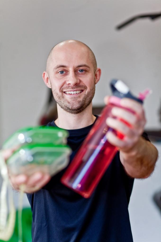 Uśmiechnięty mężczyzna z krótkimi włosami prezentuje pojemnik z jedzeniem i czerwoną butelkę na wodę, w tle fragment drążka do ćwiczeń.