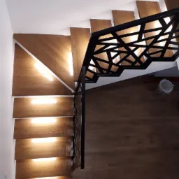 Schody drewniane z podświetleniem LED na stopniach i nowoczesną, ażurową balustradą z czarnego metalu widziane z góry.