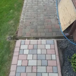 Ułożona kostka brukowa w dwóch odcieniach szarości i czerwieni, oddzielona metalowym odpływem liniowym, z widocznym trawnikiem po lewej stronie i fragmentem ściany budynku po prawej.