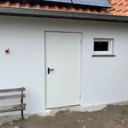 Biała ściana budynku z nową elewacją, widoczne świeżo otynkowane fragmenty, białe drzwi wejściowe z czarną klamką, małe okno i fragment dachu z dachówką oraz panelem słonecznym, obok stoi drewniana...