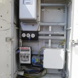 Otwarta skrzynka elektryczna z licznikiem, bezpiecznikami topikowymi, zabezpieczeniami i przewodami w kolorach: niebieskim, brązowym i zielono-żółtym. Widoczne plomby na elementach. Szara, betonowa...