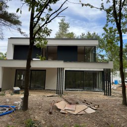 PREVGLASS - Nowoczesny dom w trakcie budowy z dużymi oknami aluminiowymi i czarnymi pionowymi elementami dekoracyjnymi na elewacji, widoczne materiały budowlane i teren wokół domu w stanie surowym.