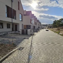 Nowe osiedle domów szeregowych z jasną elewacją i dużymi oknami, widok na brukowaną drogę i teren w trakcie zagospodarowania.