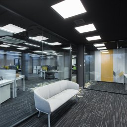 PREVGLASS - Nowoczesne biuro z przeszklonymi ścianami działowymi, widok na stanowiska pracy z roletami wewnętrznymi i geometrycznym wzorem na ścianie, oświetlenie sufitowe w kształcie kwadratów.