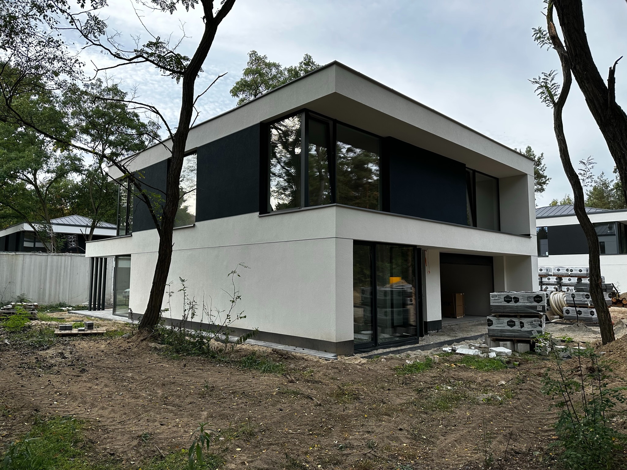 Nowoczesny dom z dużymi przeszkleniami na etapie wykończenia ogrodu, widoczne palety z materiałami budowlanymi.