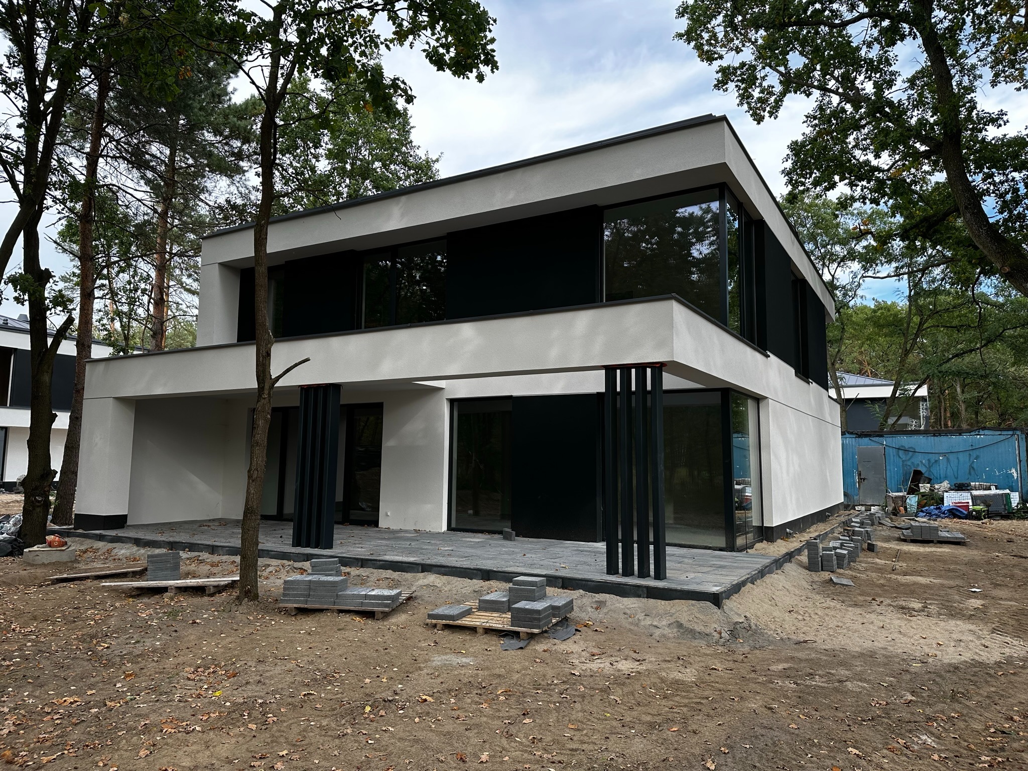 Nowoczesny dom z płaskim dachem i dużymi oknami, czarne pionowe elementy dekoracyjne, częściowo ułożony taras z szarej kostki, otoczony drzewami, teren w trakcie prac wykończeniowych.