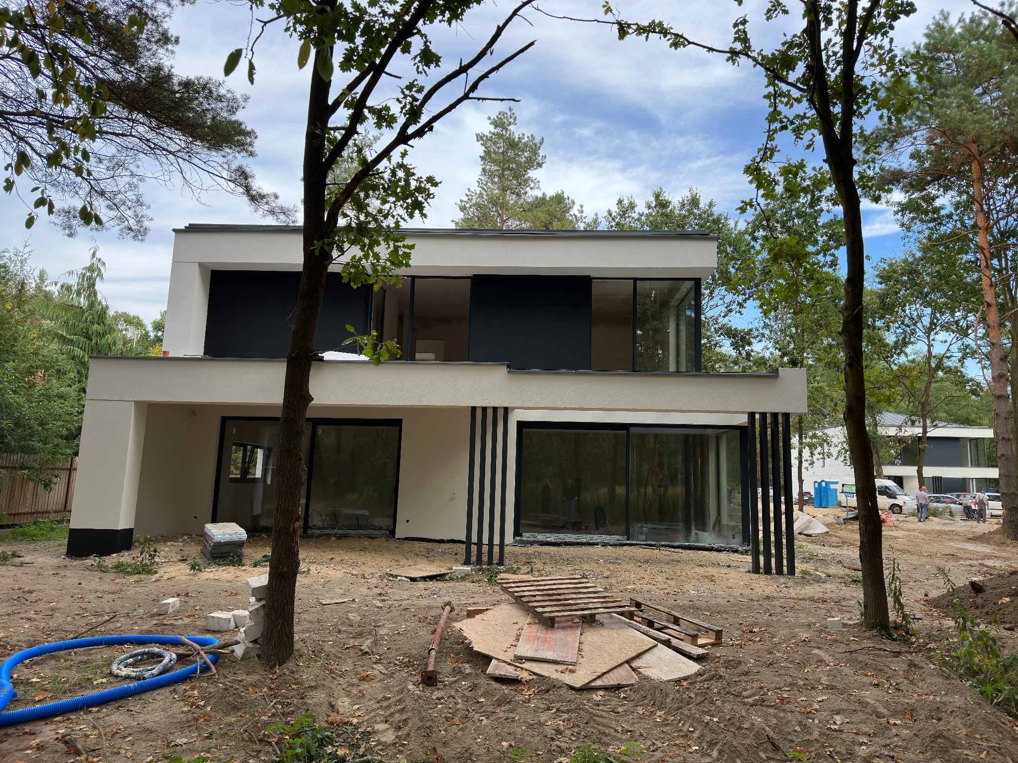 Nowoczesny dom w trakcie budowy z dużymi oknami aluminiowymi i czarnymi pionowymi elementami dekoracyjnymi na elewacji, widoczne materiały budowlane i teren wokół domu w stanie surowym.