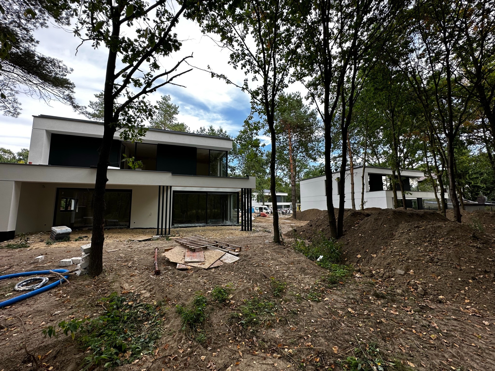 Nowoczesny dom jednorodzinny w trakcie budowy, z dużymi przeszkleniami i czarnymi ramami okiennymi, otoczony drzewami i ziemią budowlaną.