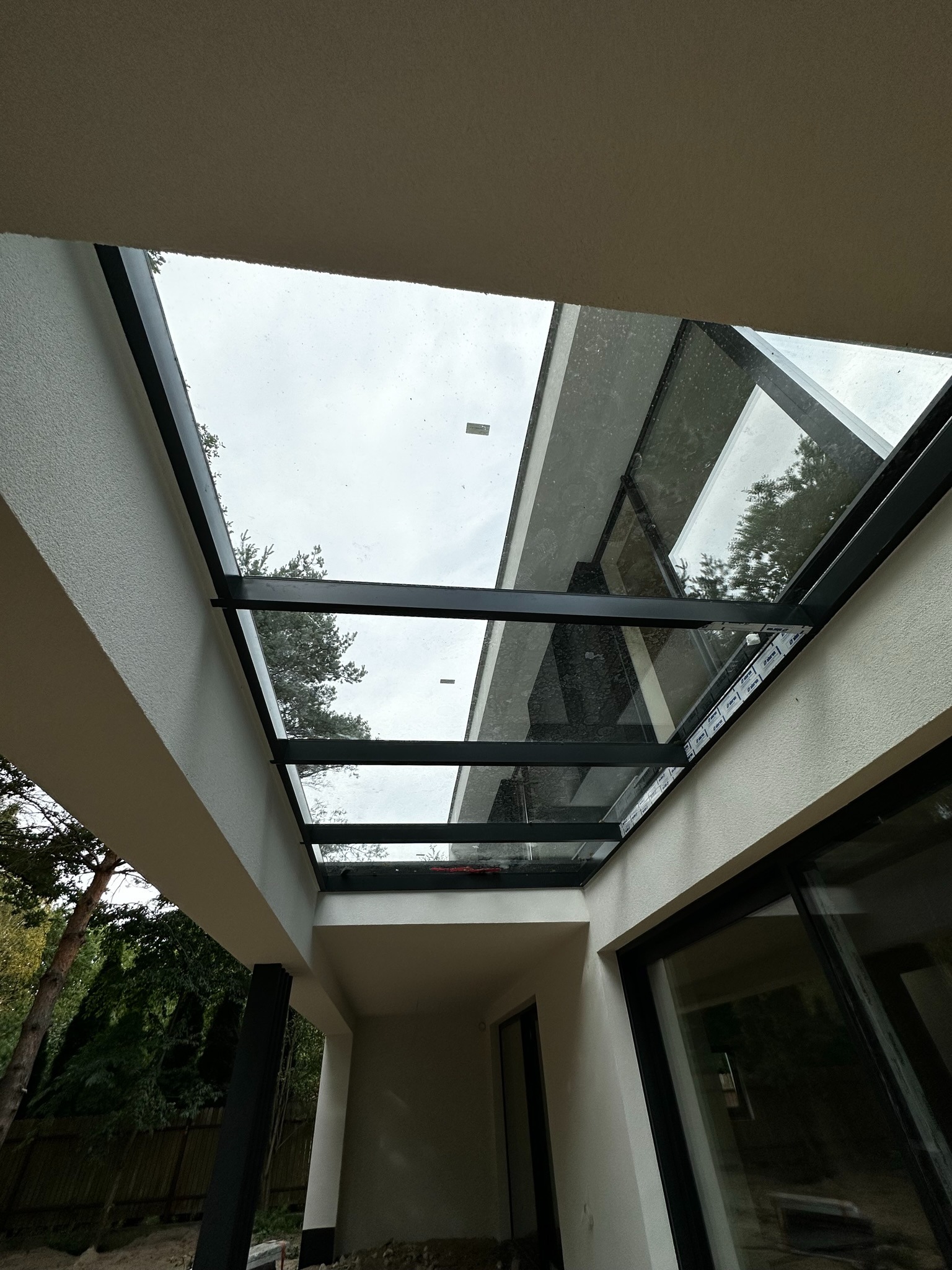 Szklane zadaszenie tarasu z czarną ramą, widok z dołu, widoczne drzewa i pochmurne niebo.