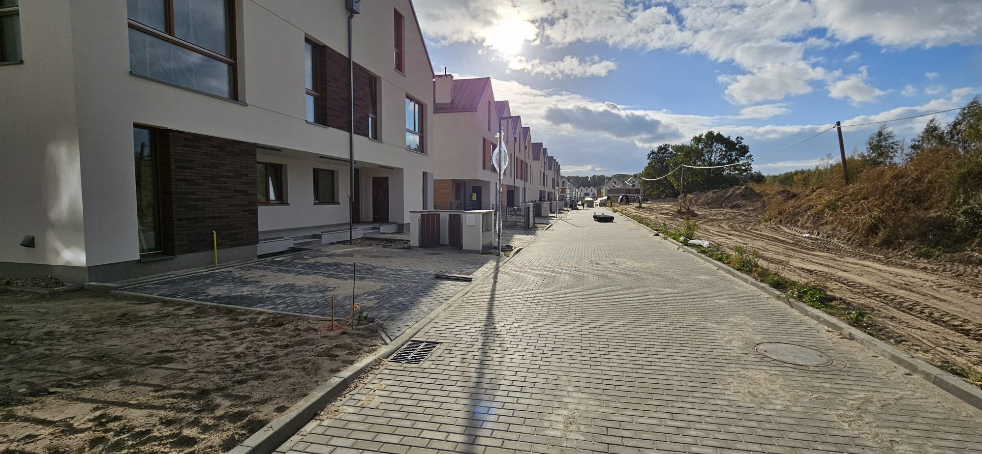 Nowe osiedle domów szeregowych z jasną elewacją i dużymi oknami, widok na brukowaną drogę i teren w trakcie zagospodarowania.