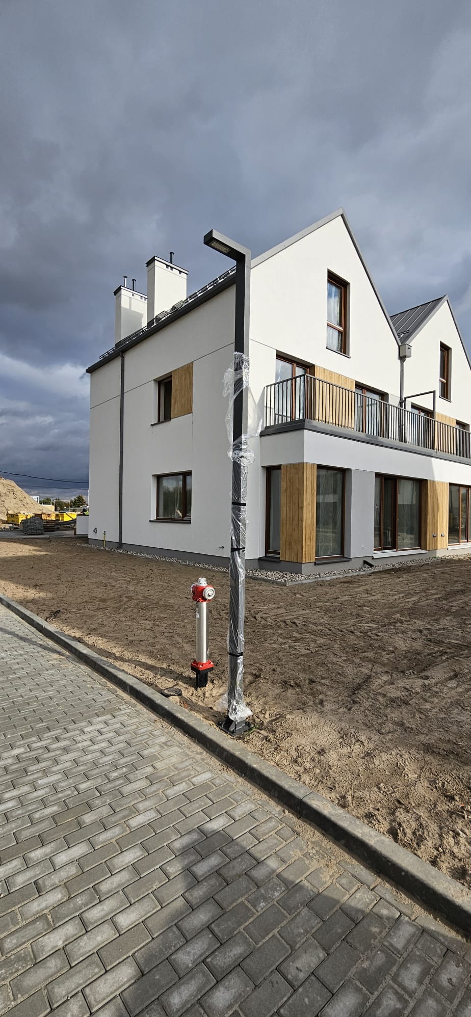 Nowoczesny dom jednorodzinny z białymi ścianami, drewnianymi akcentami i balkonem z metalową balustradą, widoczne okna z brązowymi ramami, teren wokół domu w trakcie prac wykończeniowych, szare...