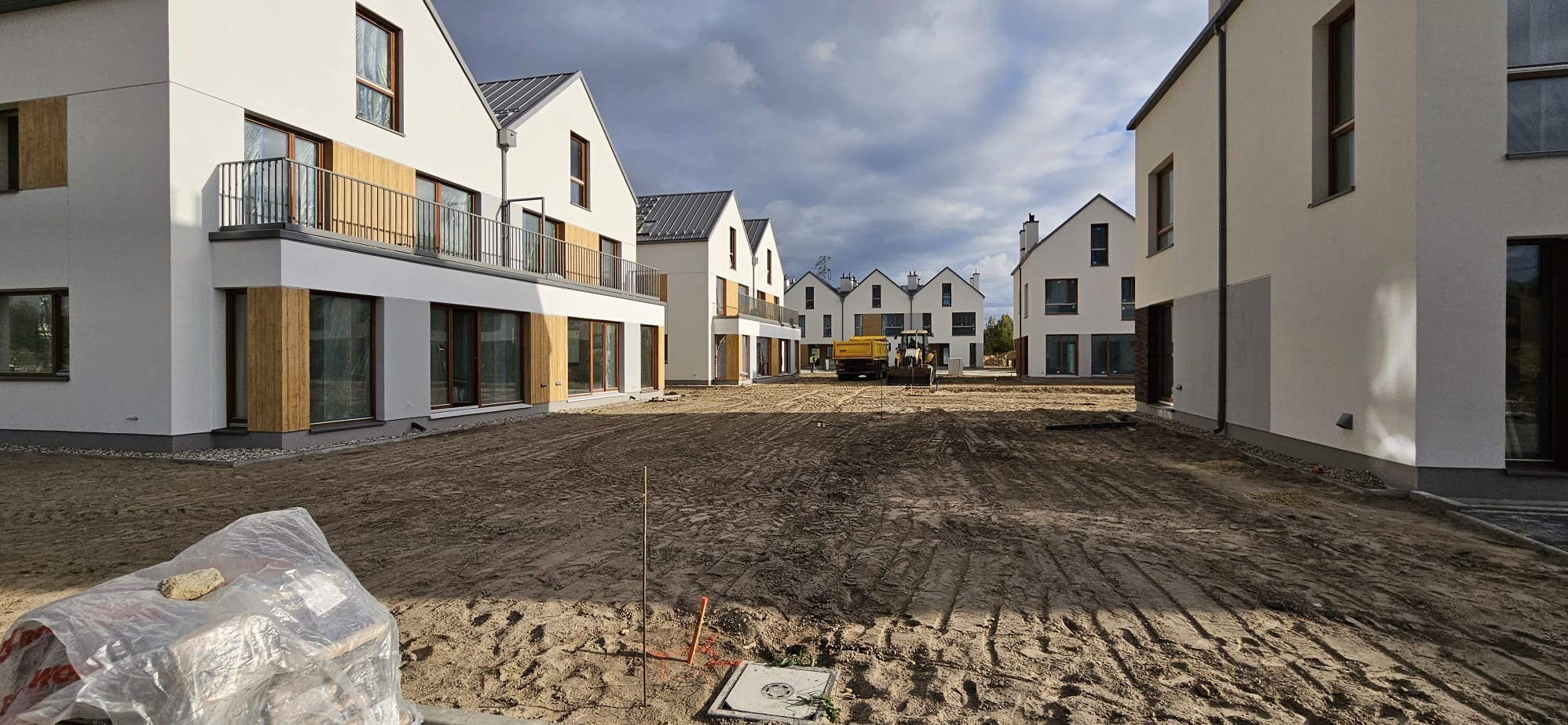 Nowoczesne osiedle domów jednorodzinnych w trakcie budowy, widoczne okna z drewnianymi elementami wykończeniowymi, teren wyrównywany przez spycharkę.