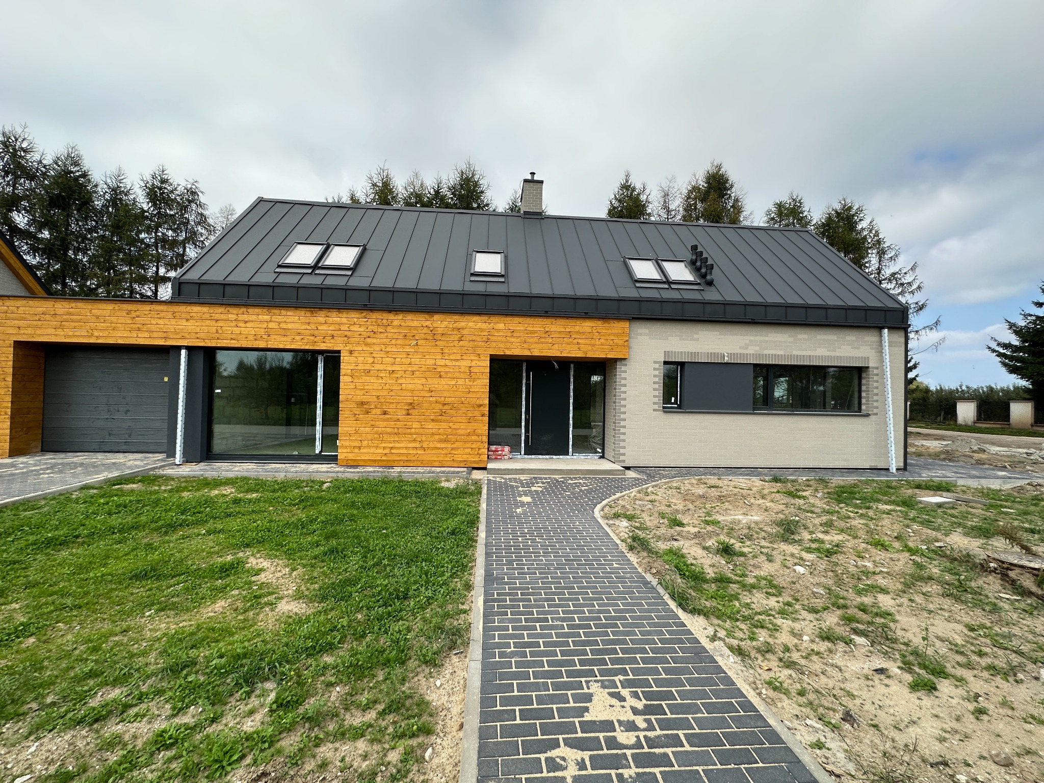 Nowoczesny dom jednorodzinny z drewnianą elewacją, szarym dachem z oknami dachowymi i brukowaną ścieżką prowadzącą do wejścia