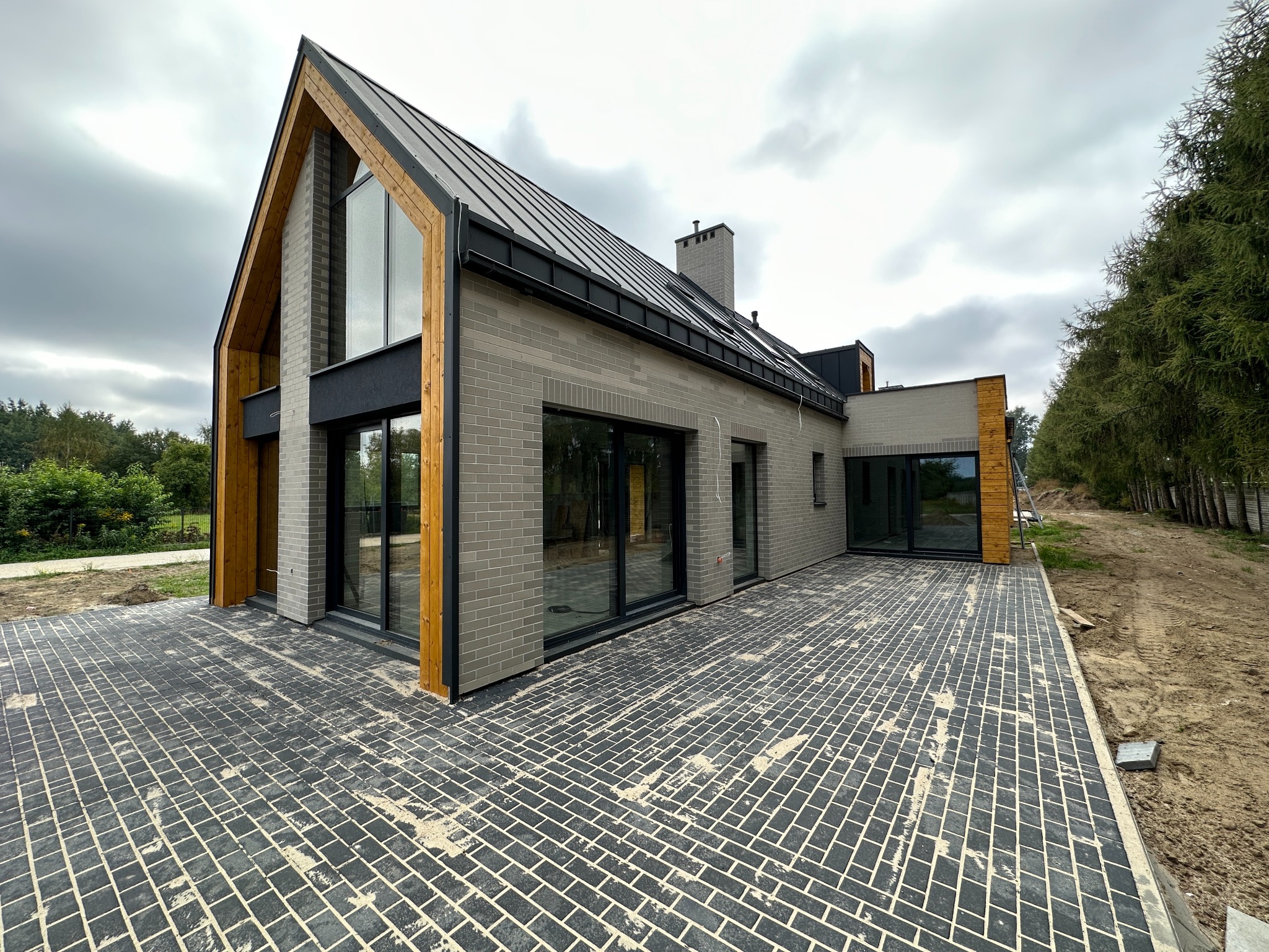 Nowoczesny dom z szarej cegły z dużymi oknami aluminiowymi i drewnianymi elementami wykończeniowymi, widok z zewnątrz na częściowo ułożony bruk.