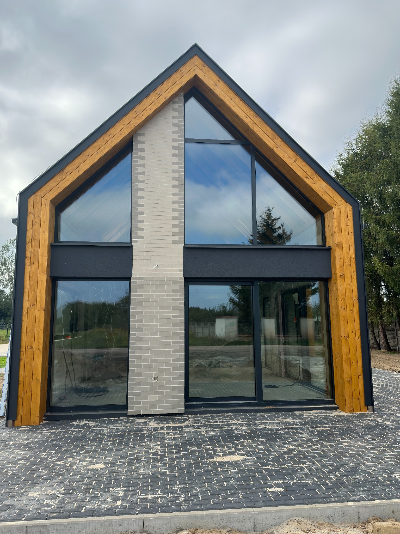 Nowoczesny dom jednorodzinny z dużymi przeszkleniami w czarnych ramach, wykończony drewnem i jasną cegłą, widok z zewnątrz na elewację frontową i brukowany podjazd.