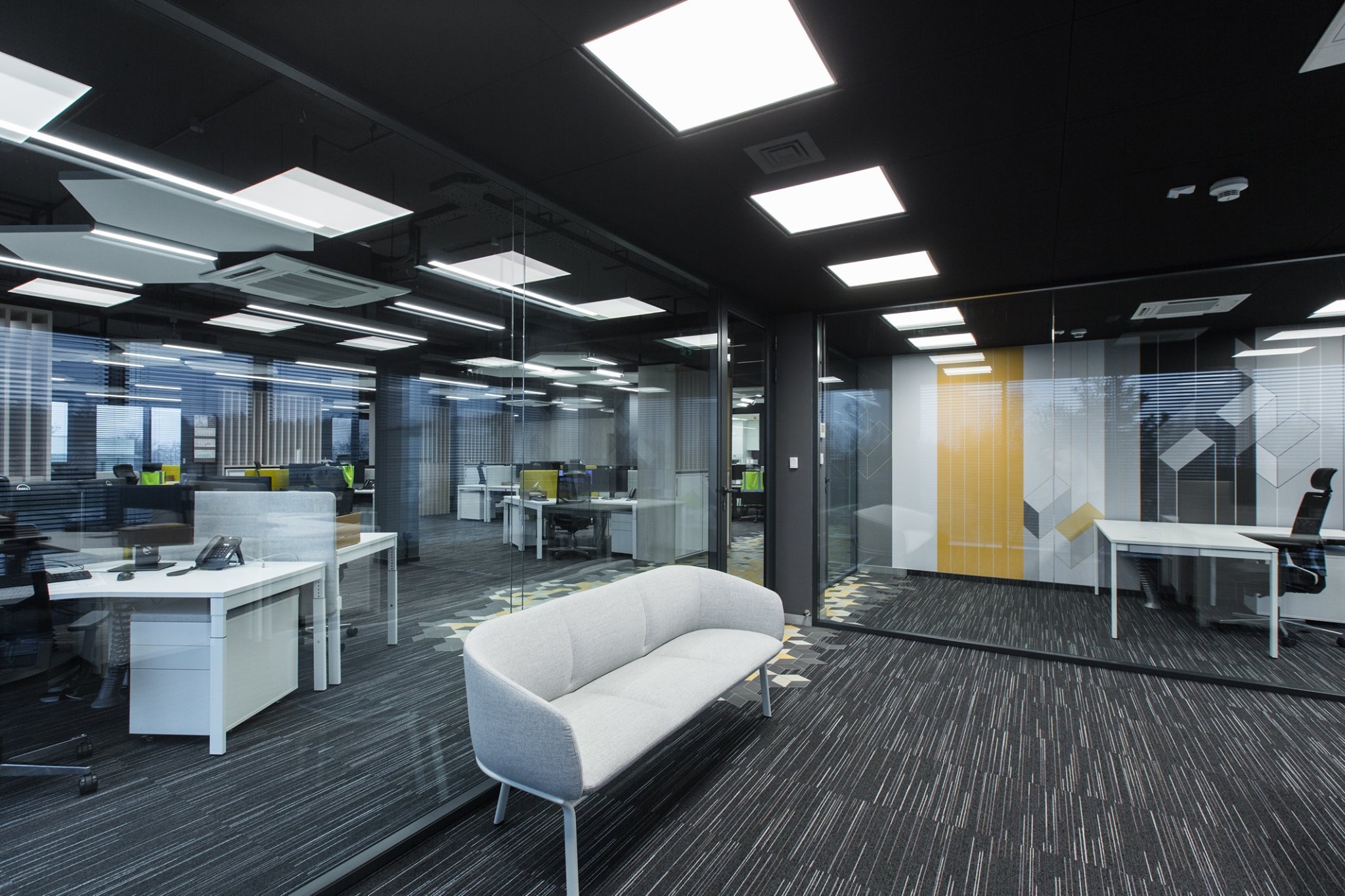Nowoczesne biuro z przeszklonymi ścianami działowymi, widok na stanowiska pracy z roletami wewnętrznymi i geometrycznym wzorem na ścianie, oświetlenie sufitowe w kształcie kwadratów.