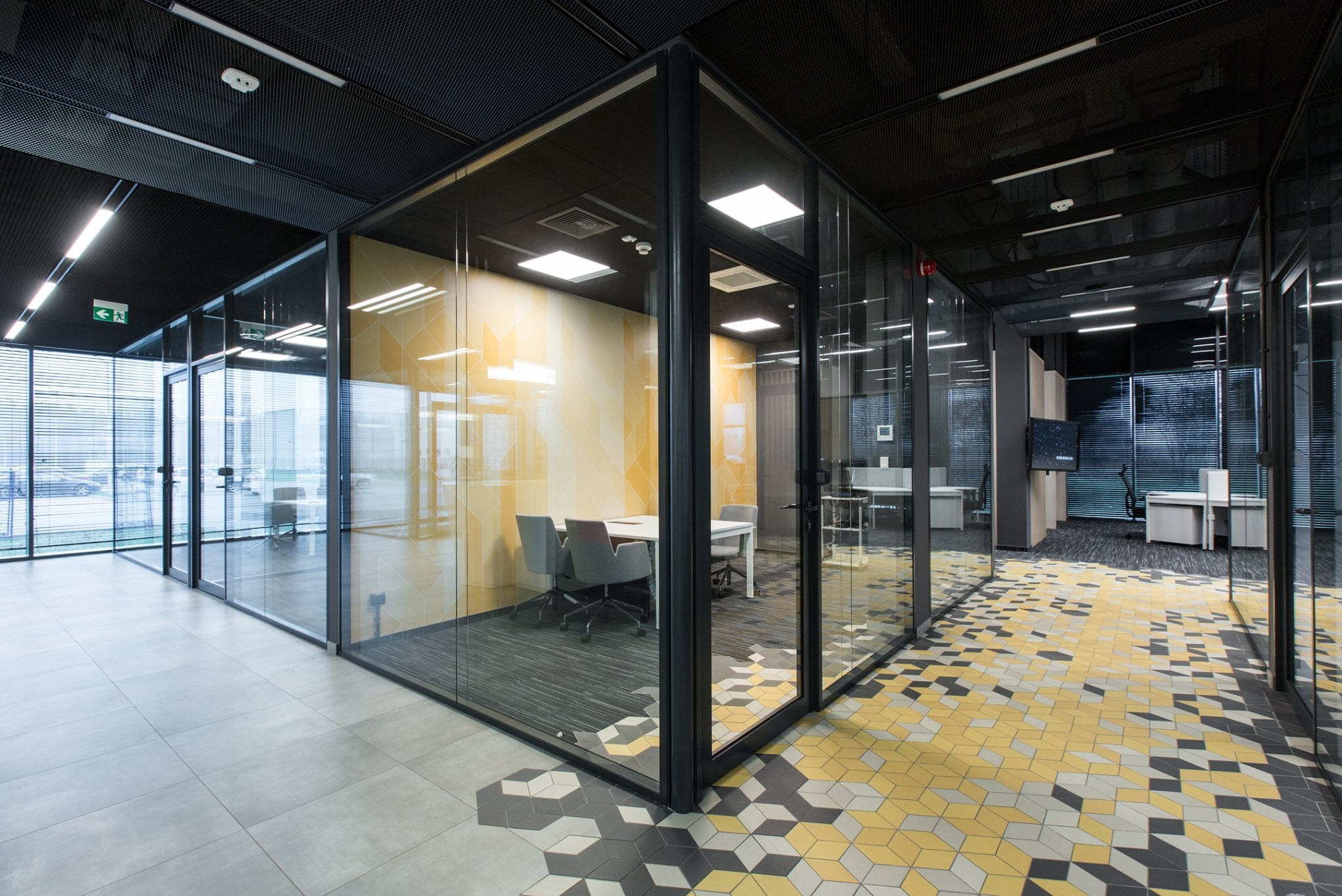 Nowoczesne biuro z przeszklonymi ścianami działowymi w czarnych ramach, widoczne żaluzje w oknach i geometryczna mozaika na podłodze.