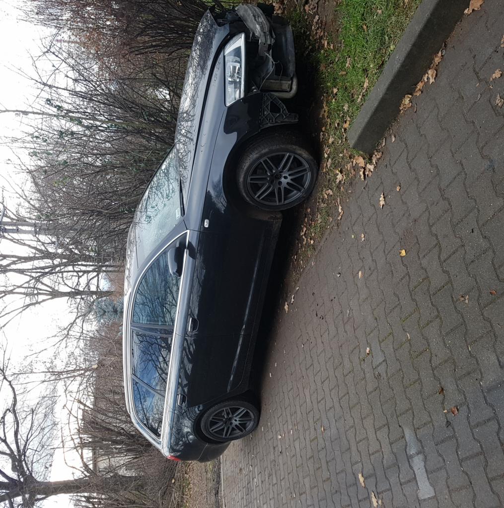 Uszkodzony przód czarnego kombi Audi na poboczu, widoczne zniszczenia karoserii i brak elementów oświetlenia.