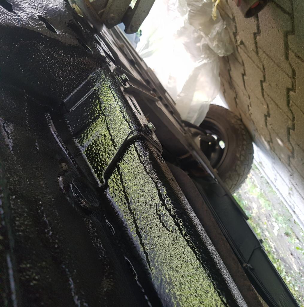 Podwozie samochodu po nałożeniu grubej warstwy czarnej powłoki antykorozyjnej, widok z dołu, z fragmentem opony w tle.