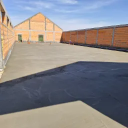 Wylany fundament pod budynek z czerwonej cegły, widok z góry. Świeży beton na dużej powierzchni, ściany w trakcie budowy. Słoneczny dzień, błękitne niebo.