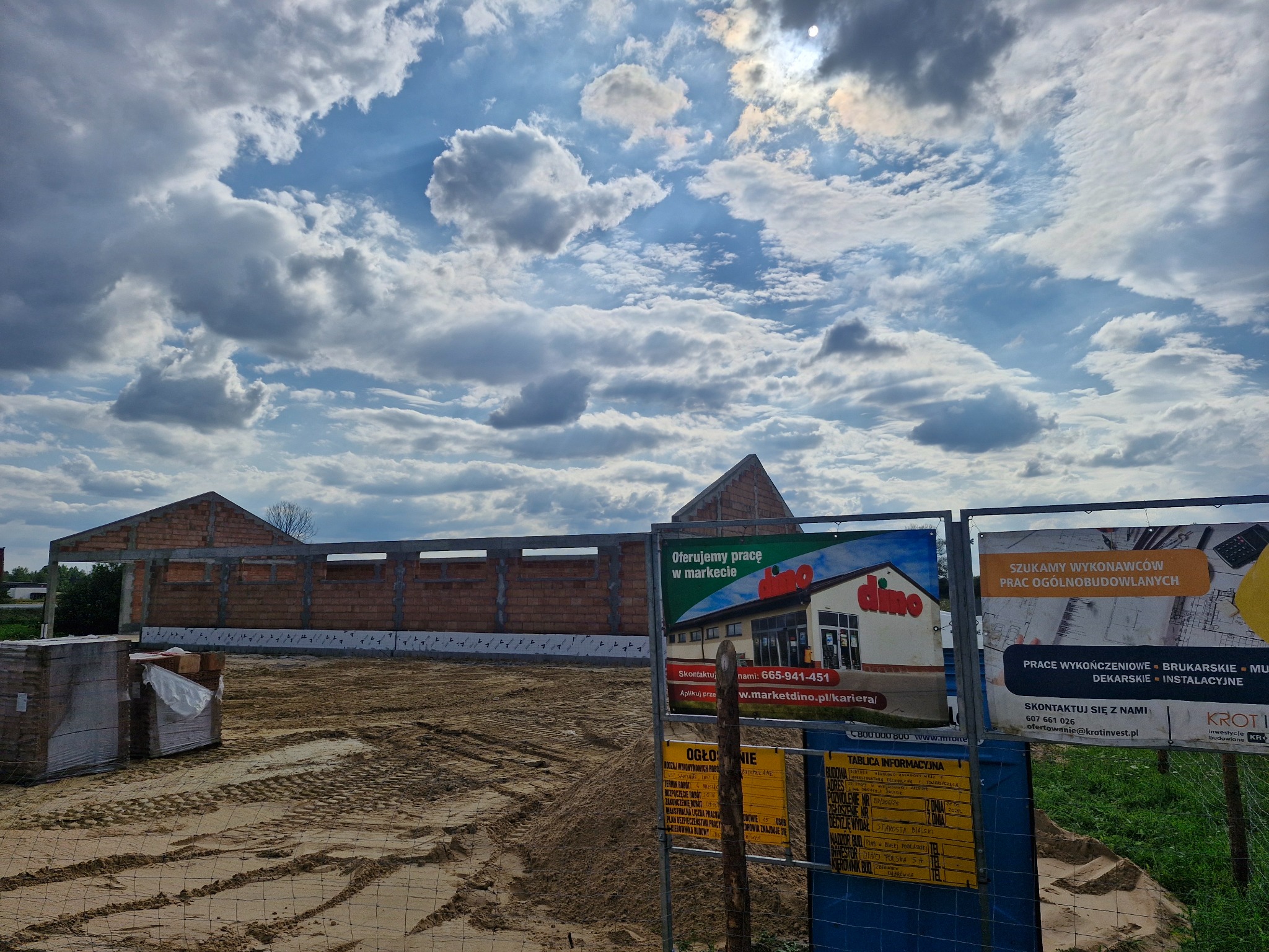 Wznoszenie murów budynku z czerwonej cegły, widoczne reklamy na budowie. Niebo z chmurami w tle. Etap budowy obiektu usługowego z cegły.