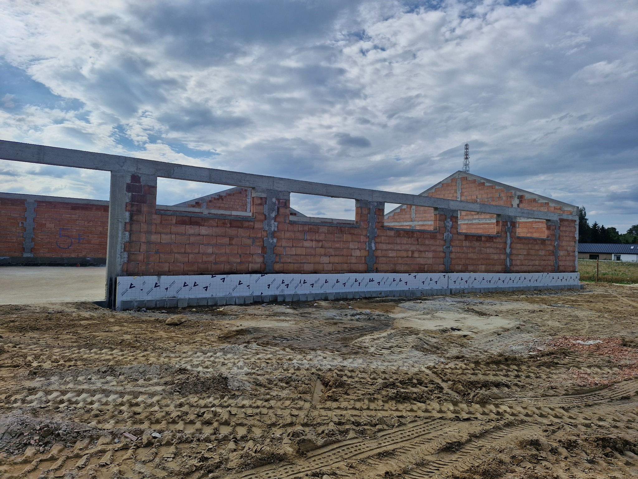 Wznoszenie ścian z czerwonej cegły na fundamencie z izolacją przeciwwilgociową. Widoczne betonowe nadproża. Tło: pochmurne niebo i fragment krajobrazu.