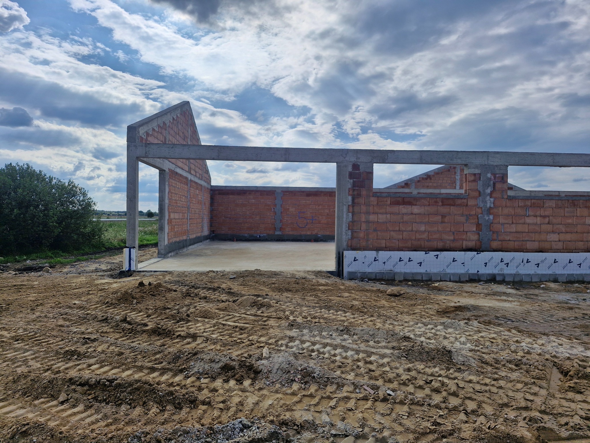 Surowy stan budynku w trakcie budowy: murowane ściany z czerwonej cegły, betonowe słupy i strop, widoczne fundamenty z izolacją, teren budowy z ziemią.