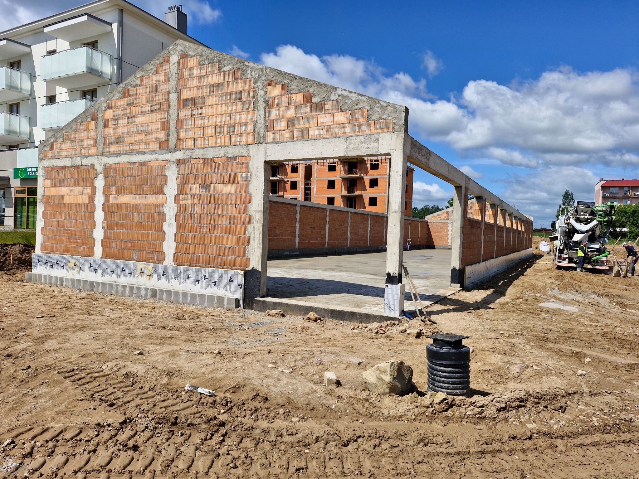 Surowy stan domu murowanego z betonu komórkowego, widoczne pustaki i betonowe słupy, w tle budynek mieszkalny i betoniarka. Teren budowy w słoneczny dzień.