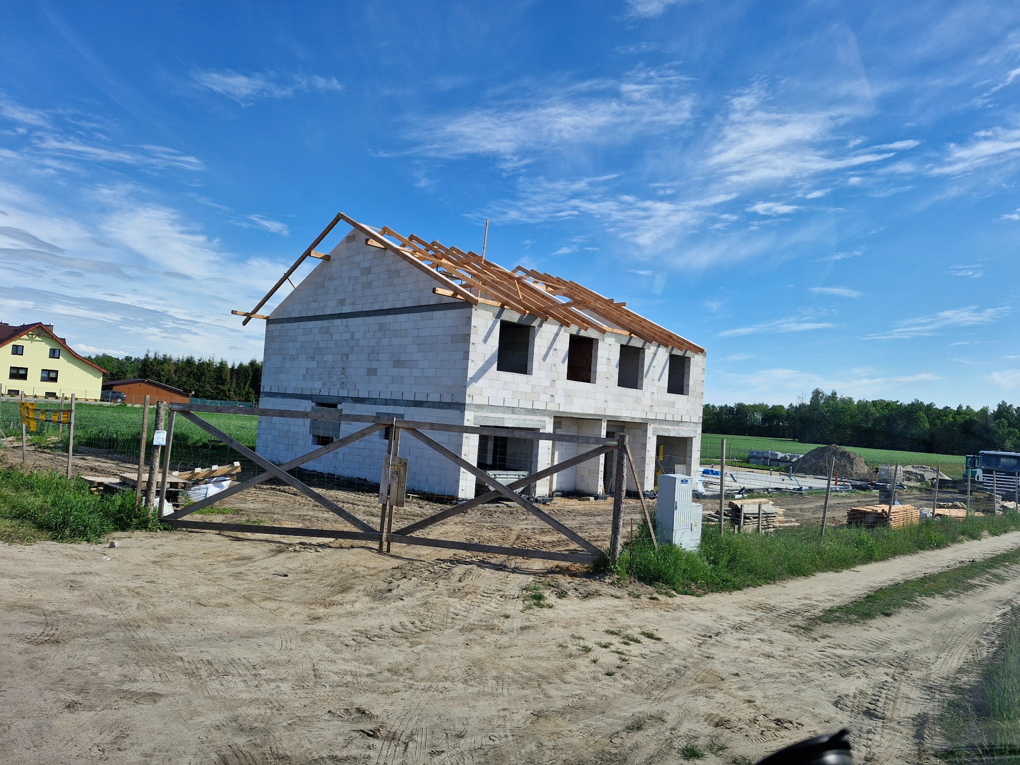 Dwukondygnacyjny dom w stanie surowym otwartym z drewnianą konstrukcją dachu na tle błękitnego nieba z obłokami, ogrodzony drewnianym płotem.