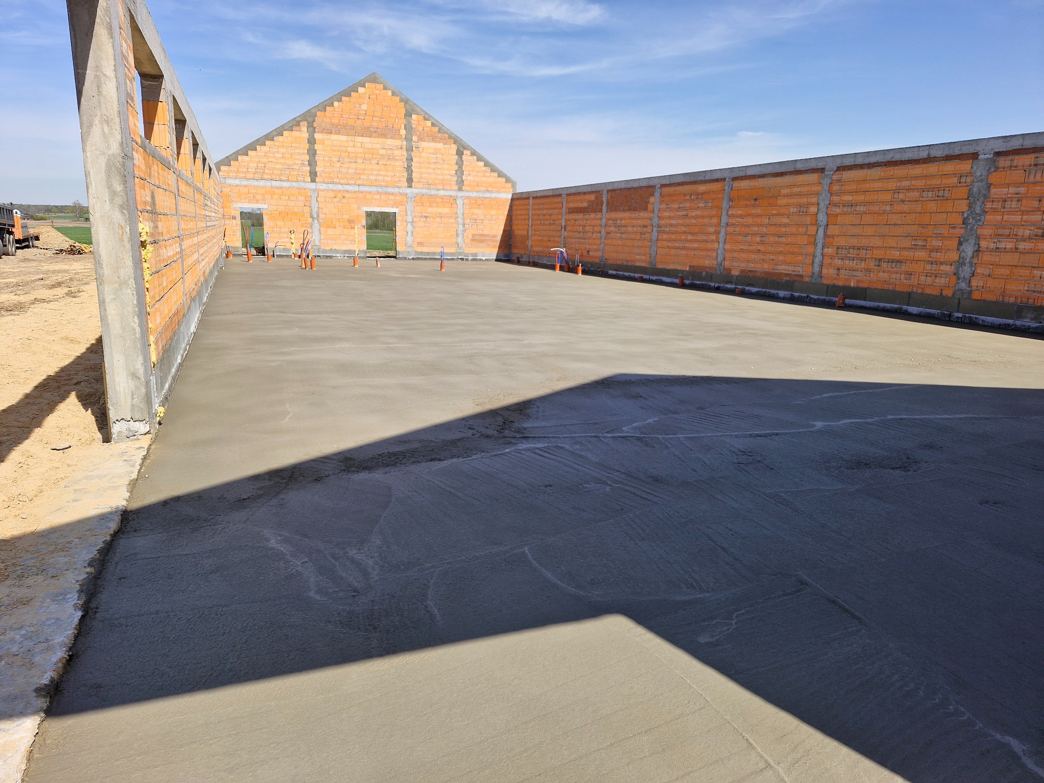 Wylany fundament pod budynek z czerwonej cegły, widok z góry. Świeży beton na dużej powierzchni, ściany w trakcie budowy. Słoneczny dzień, błękitne niebo.