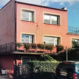 Dwupiętrowy dom w odcieniach różu z kremowymi obramowaniami okien, balkonem ozdobionym kwiatami i anteną na elewacji, widziany z poziomu ulicy.