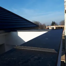 Fragment dachu z czarną dachówką ceramiczną i obróbkami blacharskimi w kolorze antracytu, widok z rusztowania na budowę domu szkieletowego w tle.