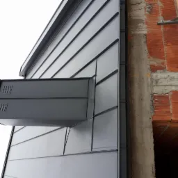 Szara, metalowa elewacja budynku w trakcie budowy z widoczną wentylacją i fragmentem muru z czerwonej cegły.