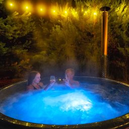 Sauna Ogrodowa BIBERHAUS - Para w balii ogrodowej z hydromasażem, podświetlonej na niebiesko, w otoczeniu iglaków i girlandy światełek, trzymająca kieliszki z winem.