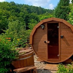 Sauna Ogrodowa BIBERHAUS - Drewniana beczka sauny z przeszklonymi drzwiami i lampą na zewnątrz, otoczona zielenią ogrodu. Obok stoi drewniana balia.