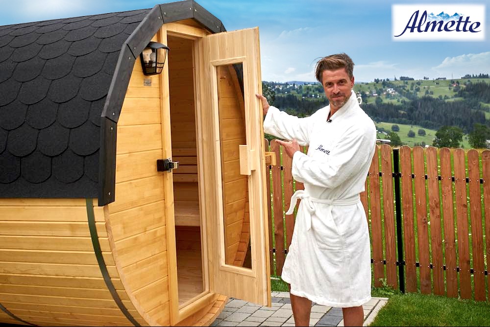 Beczkowa sauna z otwartymi drzwiami, prezentowana przez mężczyznę w szlafroku na tle górskiego krajobrazu.