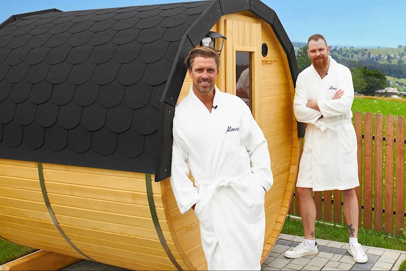Drewniana beczka z czarnym dachem krytym gontem, pełniąca funkcję sauny, prezentowana przez dwóch mężczyzn w białych szlafrokach z logo 'Almeti' na tle zielonego krajobrazu z drewnianym płotem.